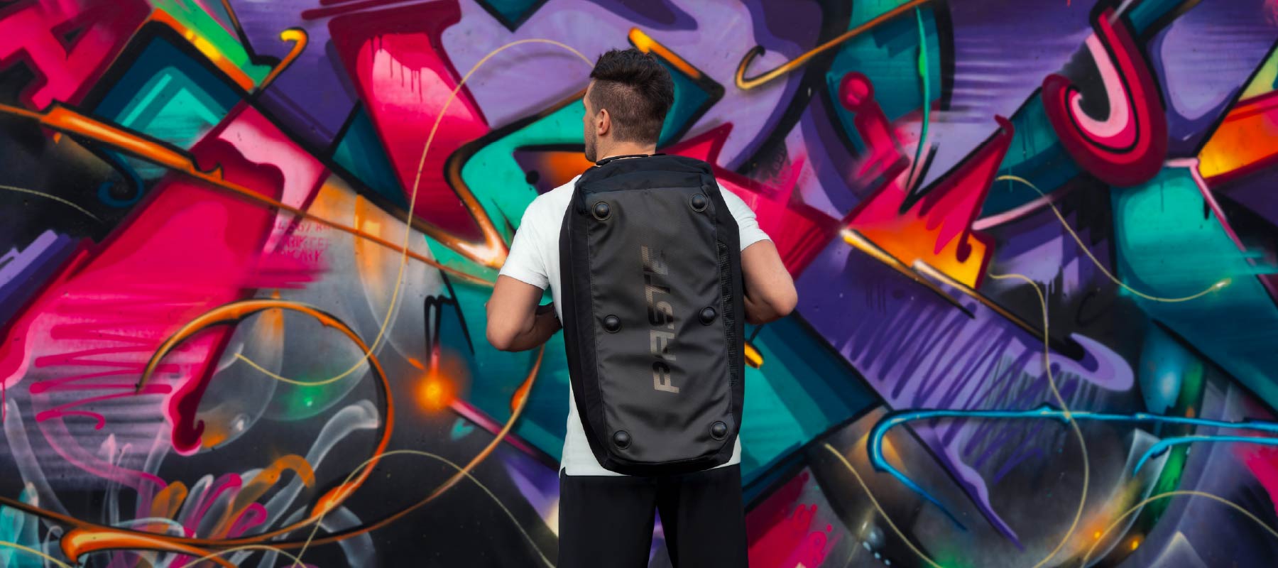 Model trägt die PASTE ALL IN PRO Sporttasche als Rucksack vor einer farbigen Graffitiwand.